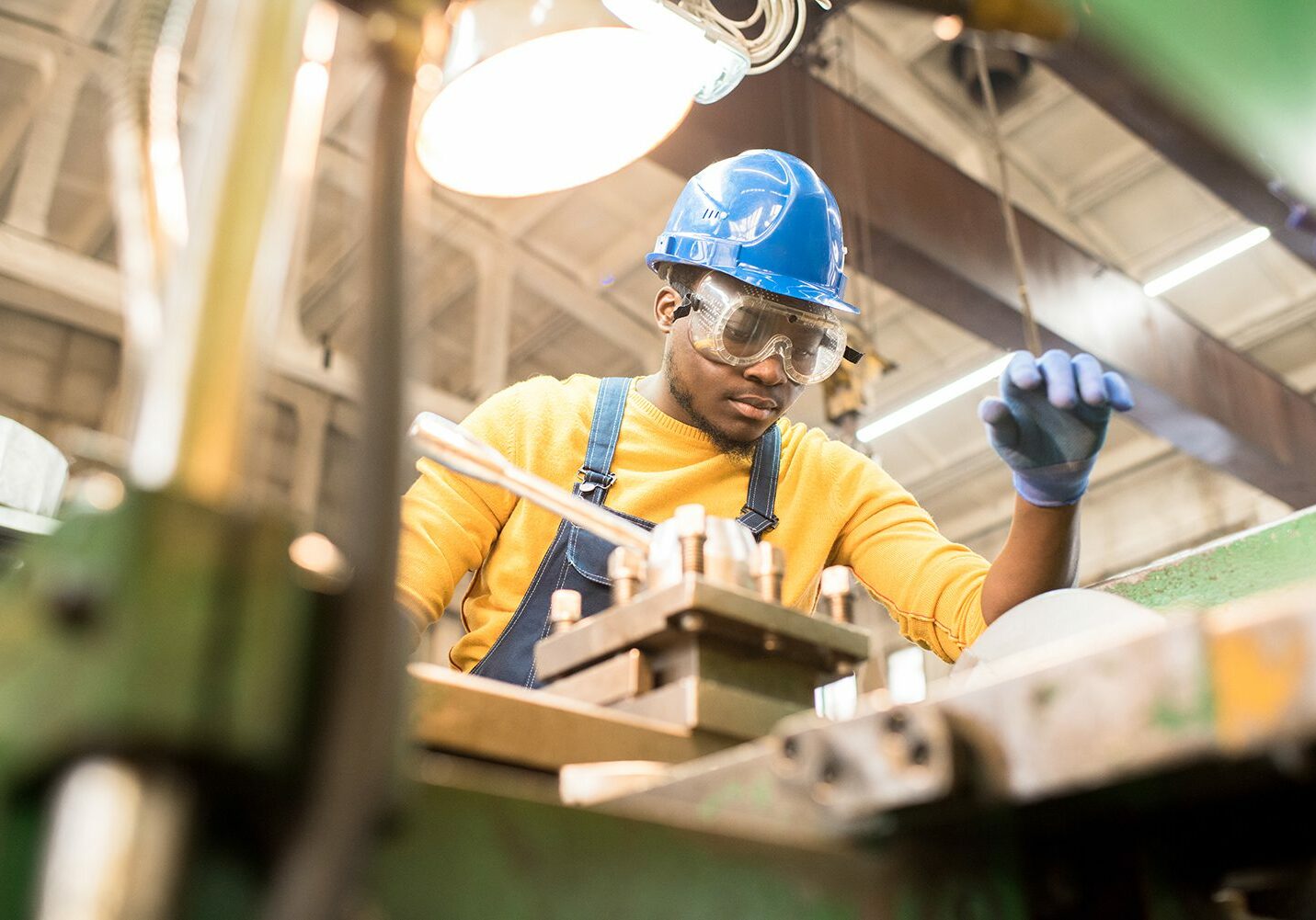 Serious busy young black factory engineer in hardhat and safety goggles examining milling lathe and repairing it while working at production plant