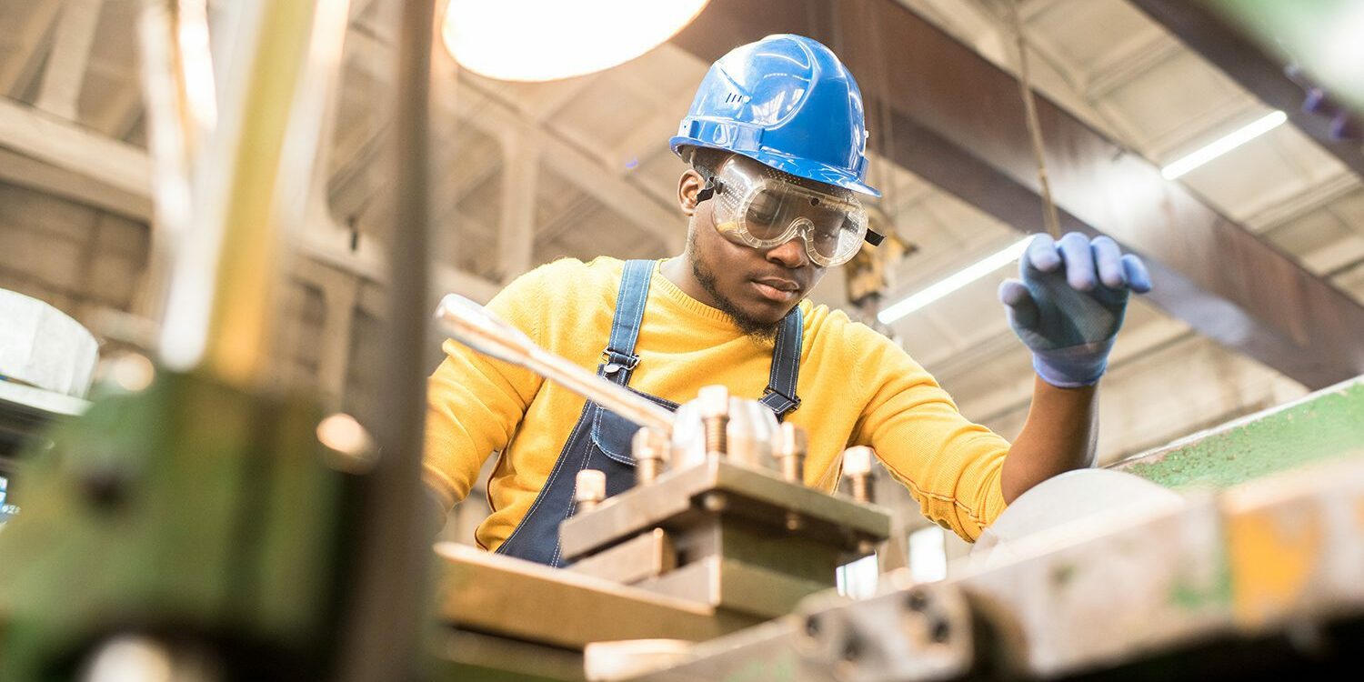 Serious busy young black factory engineer in hardhat and safety goggles examining milling lathe and repairing it while working at production plant