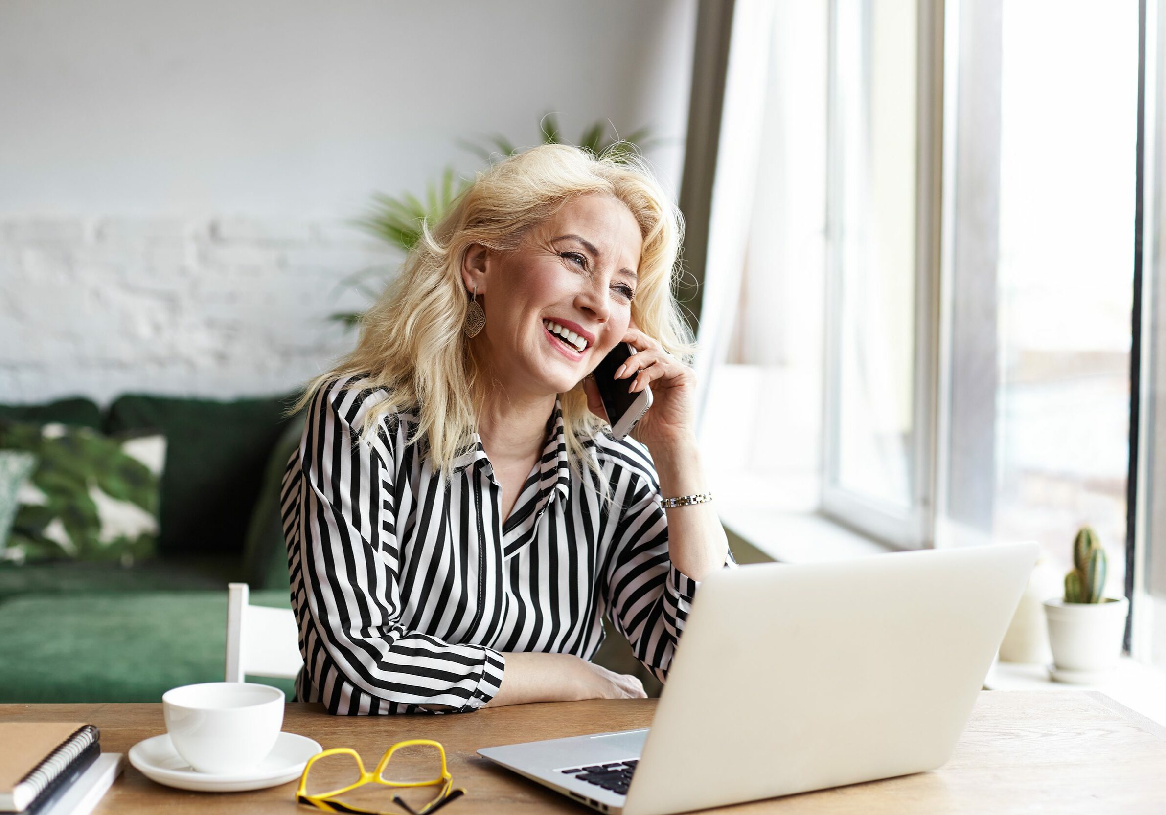 Attractive successful elderly businesswoman in striped blouse working in modern office, making phone call to potential client, having nice conversation, sitting at desk in front of open laptop