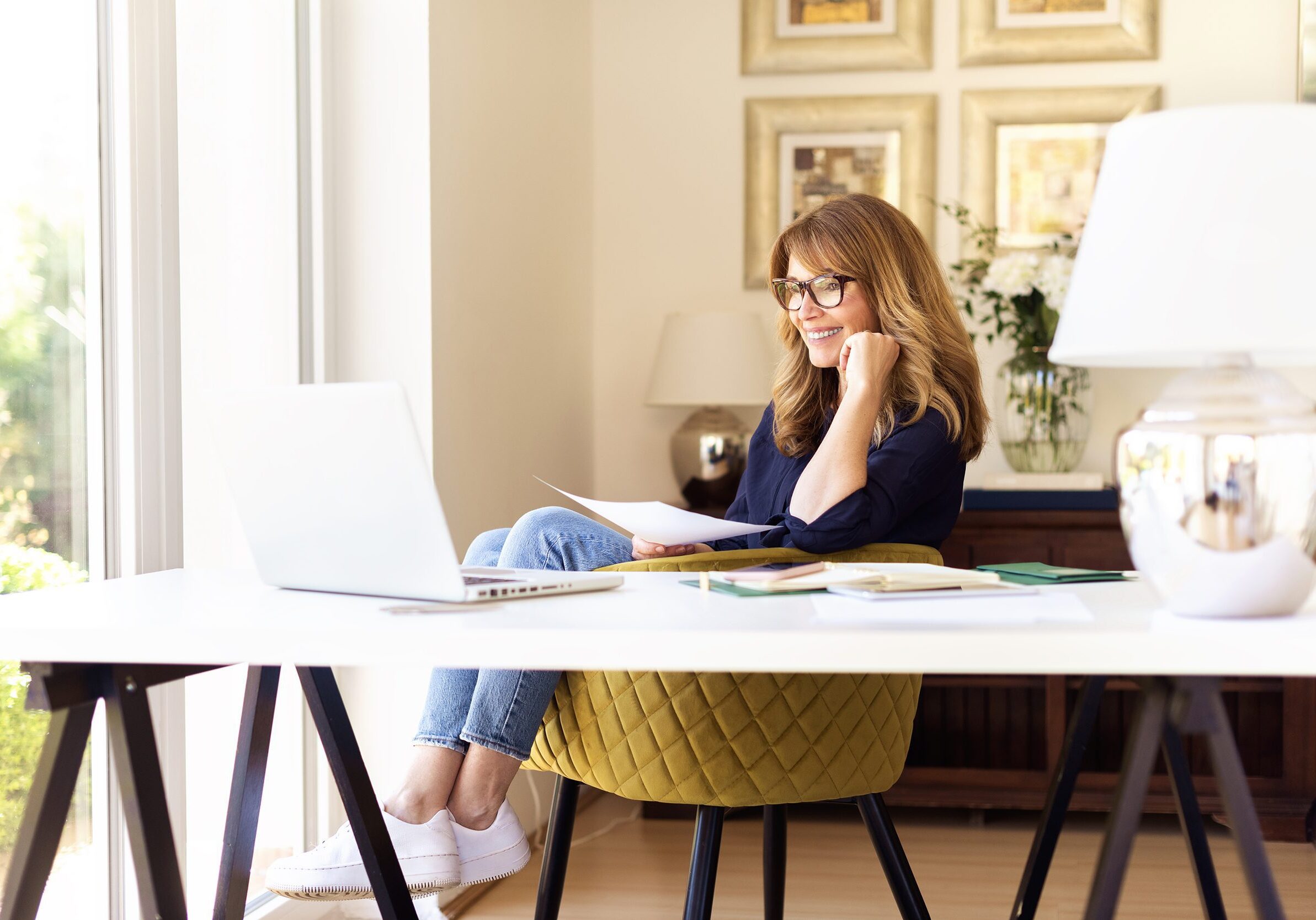 Attractive mature woman sitting at desk and using laptop while w Shot of happy smiling businesswoman using her laptop while working at home. Home office.